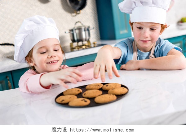 两个小孩在拿饼干愉快的小孩子在厨师帽子吃可口曲奇饼在厨房